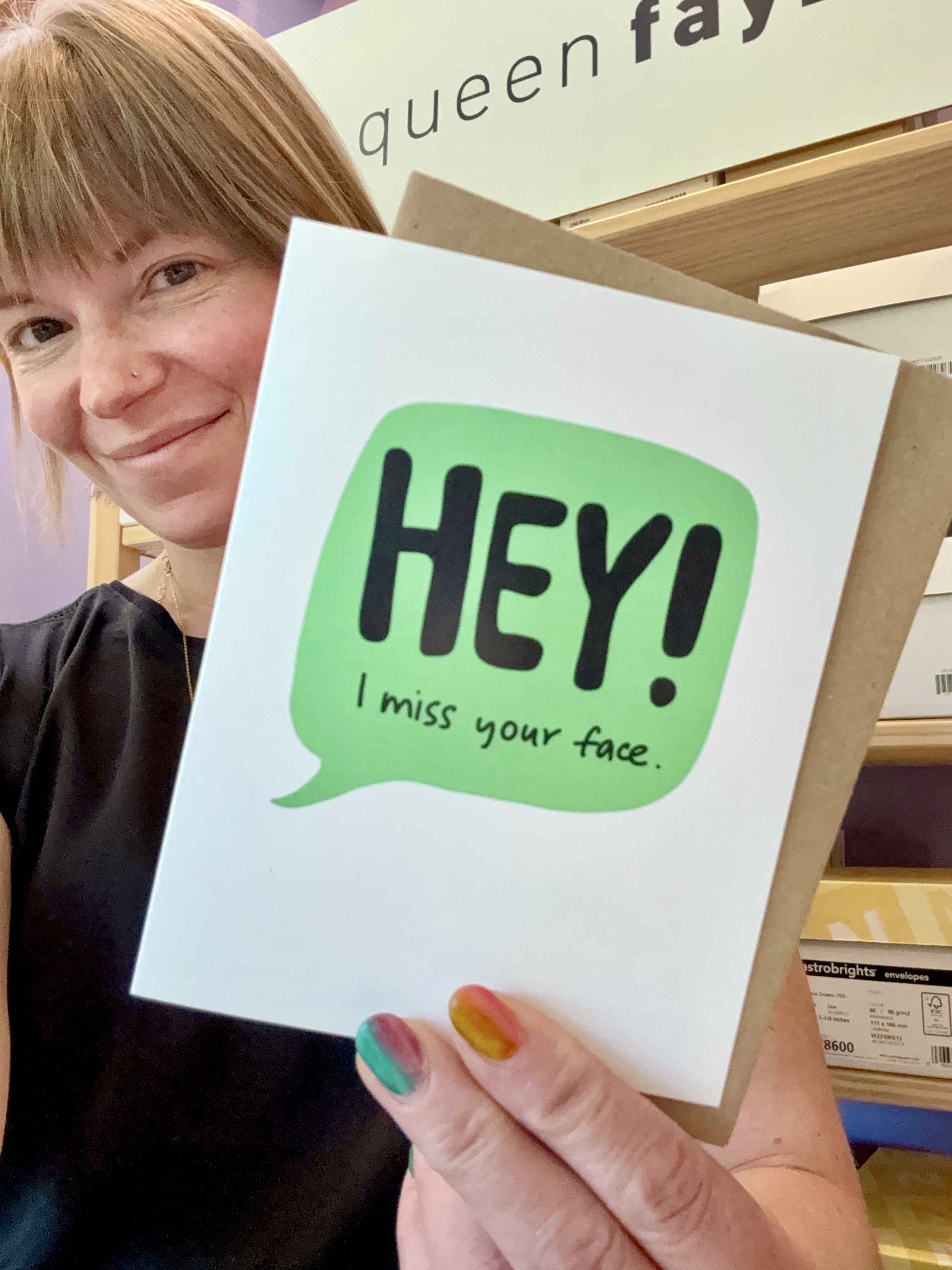 a white woman holding a friendship greeting card up to the camera. the card is white with a green speech bubble and the words, "HEY! I miss your face."