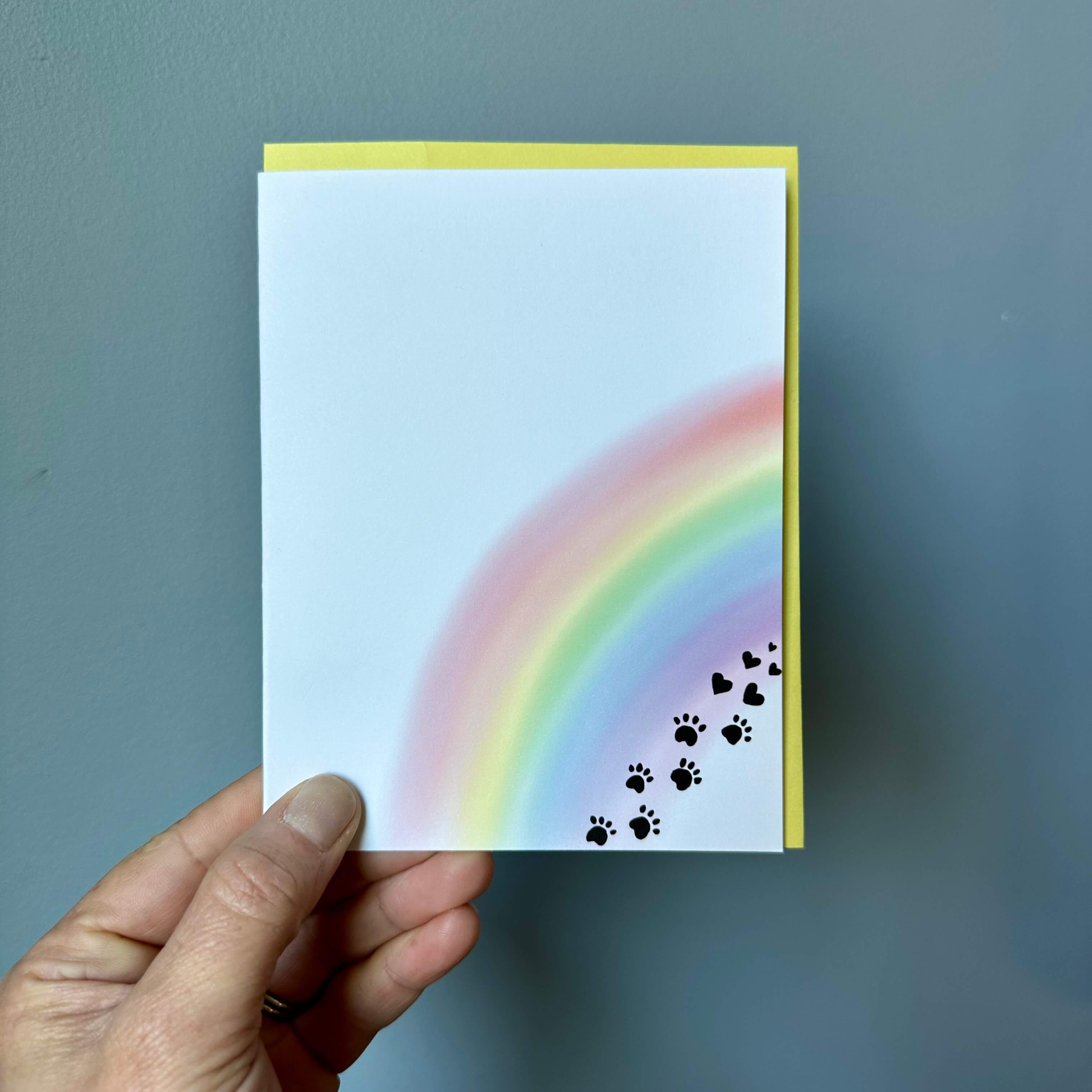 hand holding a pet loss sympathy card against a gray background. the card has a watercolor rainbow with black paw prints that transition to hearts.