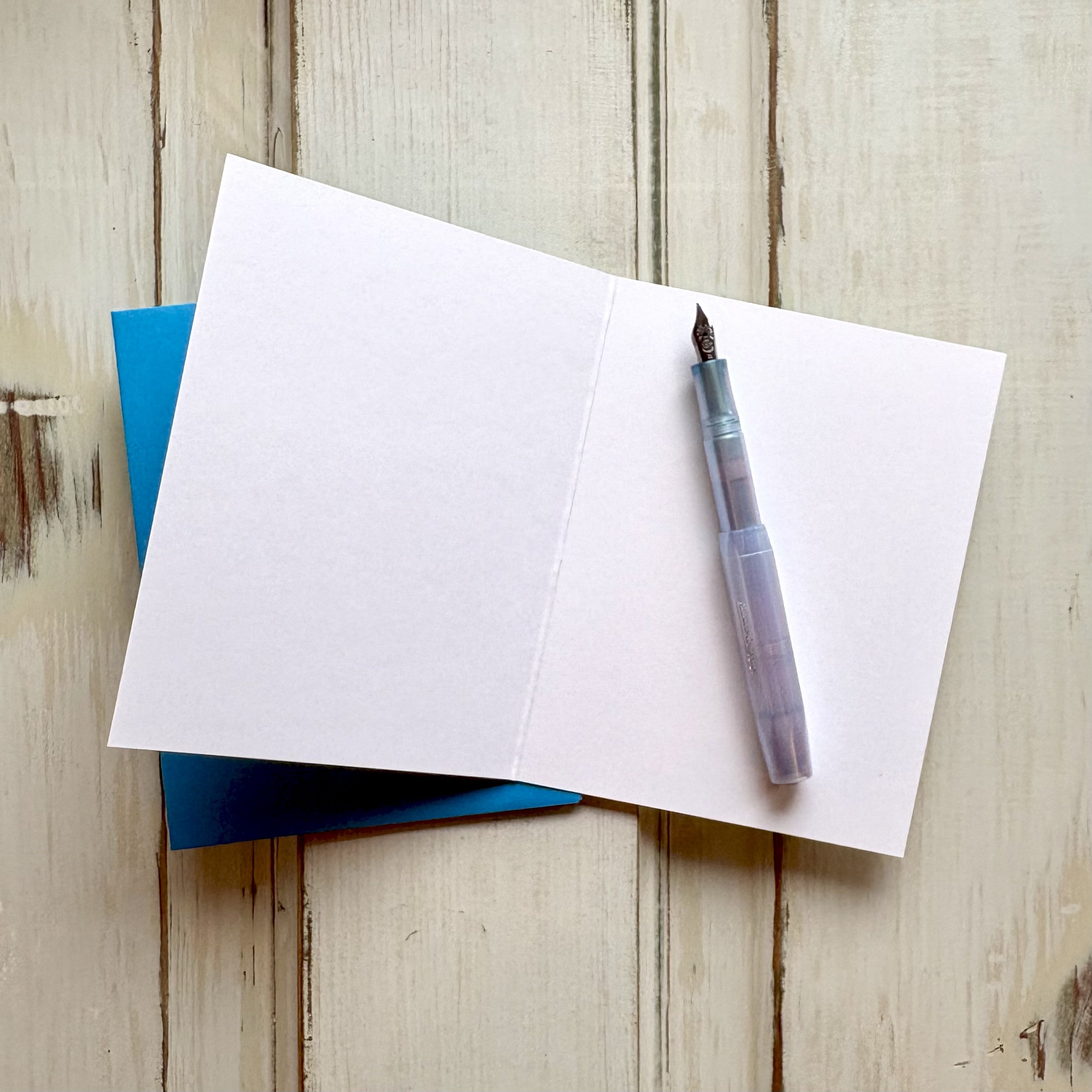Open blank greeting card with a fountain pen and blue envelope, on a wooden desk.