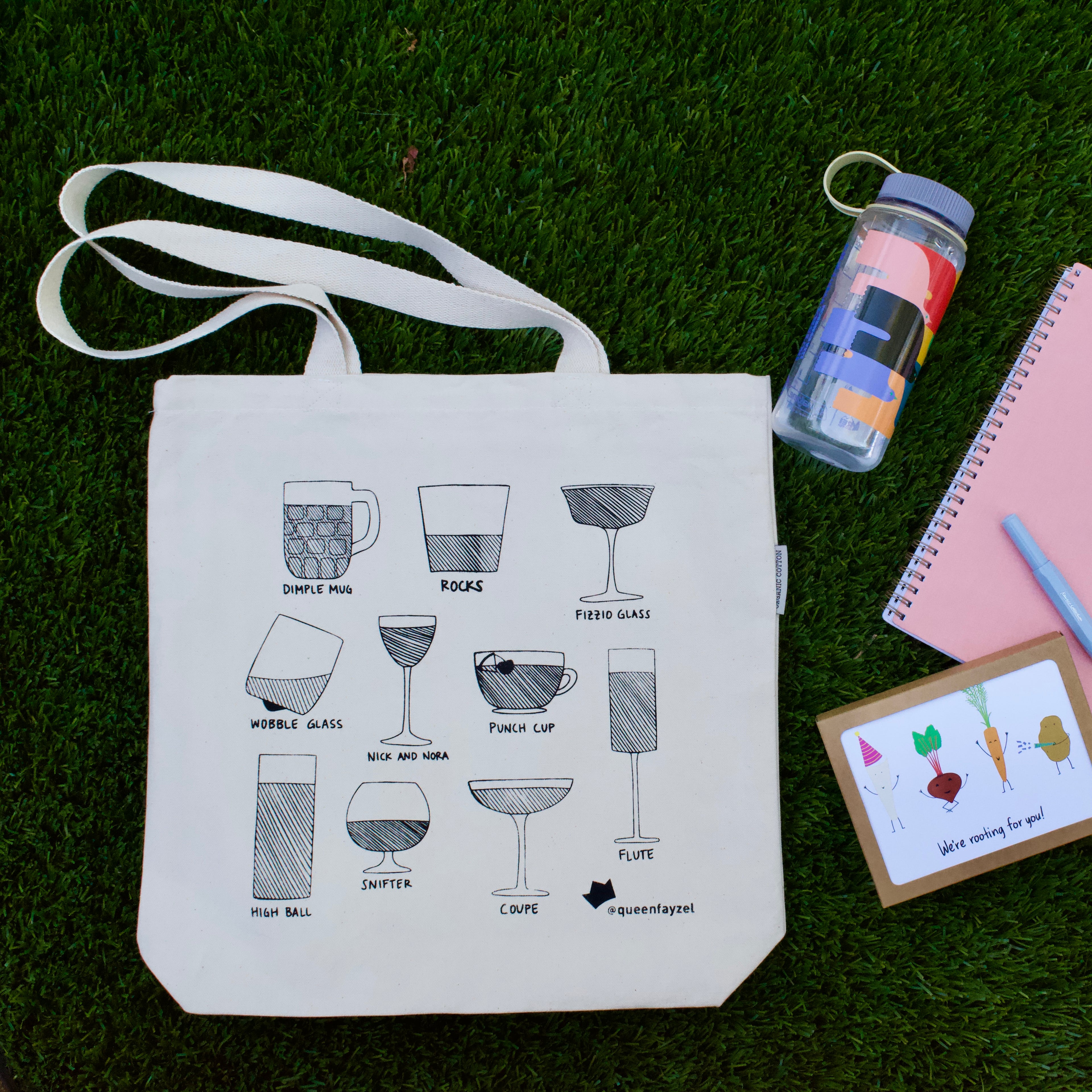 canvas tote bag with drinkware design arranged in grass with a water bottle, notebook, pen, and set of greeting cards