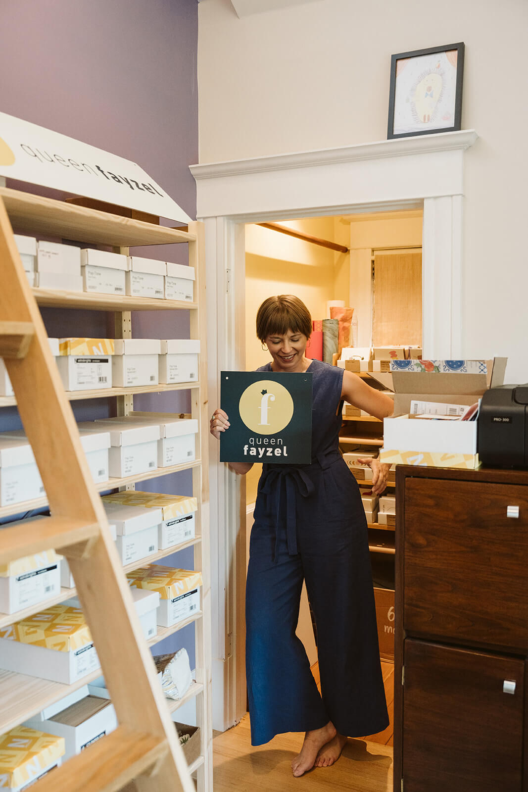 greeting card designer Faye holding a Queen Fayzel sign. there is a shelf of envelopes is in the foreground.