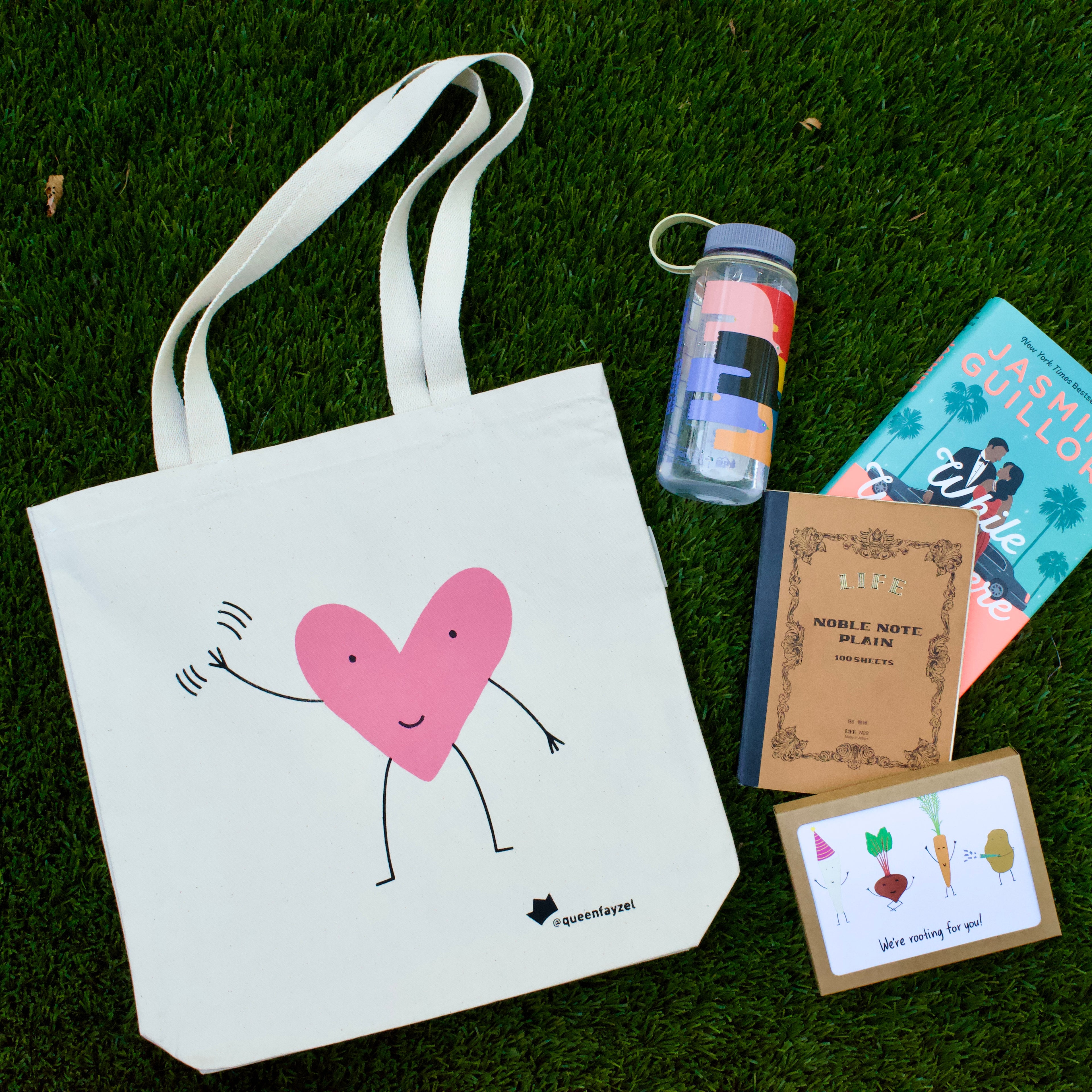 cute tote bag with waving heart design laid out on grass with a water bottle, journal, book, and set of greeting cards