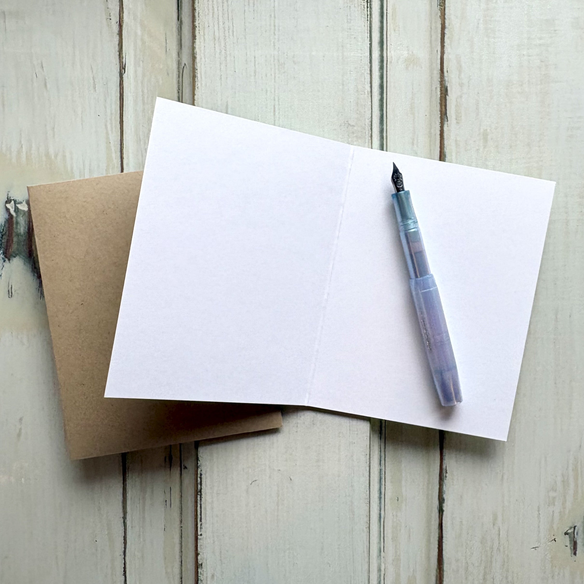 Fountain pen and blank card with a tan envelope on a wooden surface.