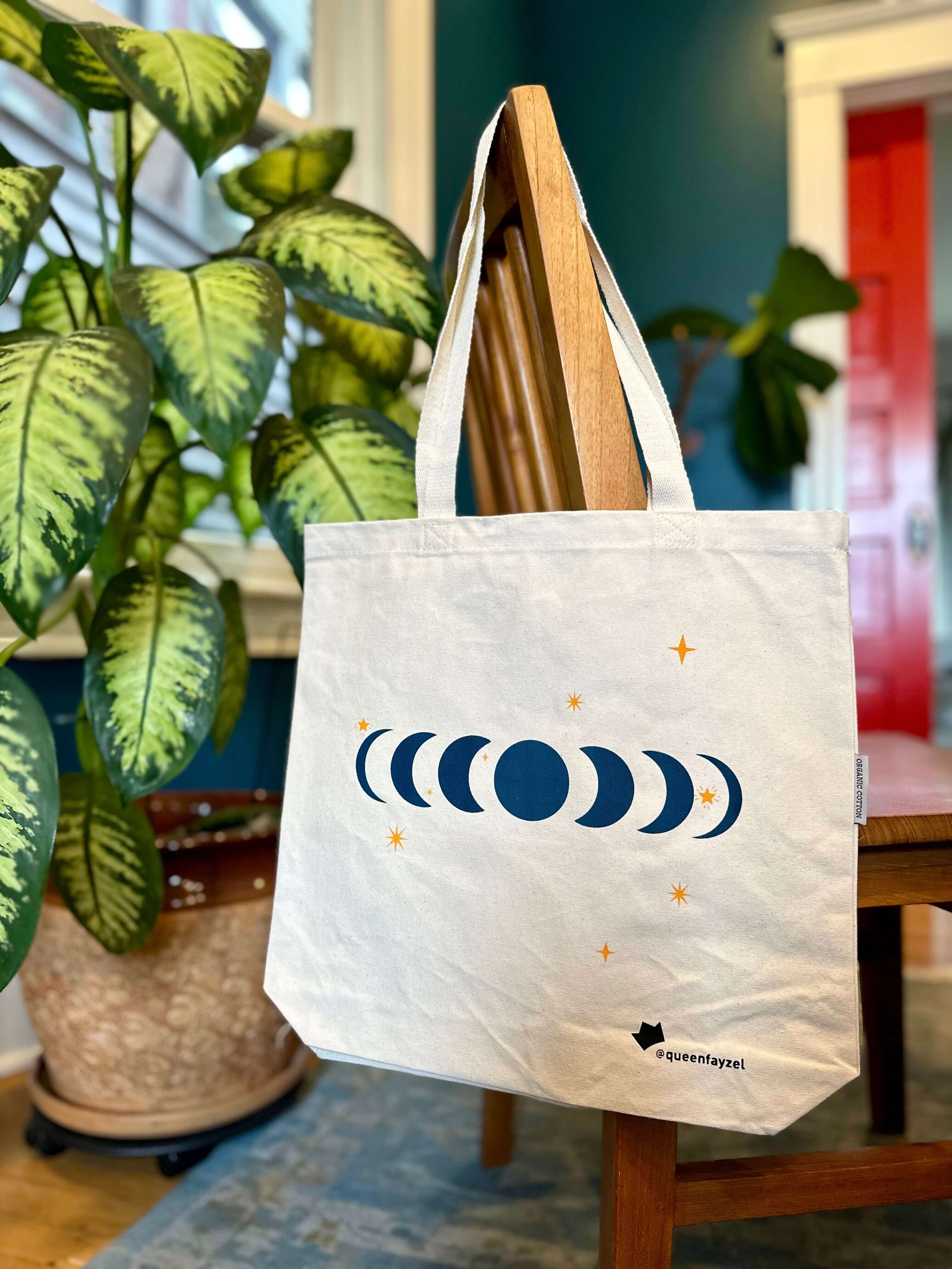 cotton tote bag with moon phase design hanging on the back of a wooden chair with a plant and blue wall in the background