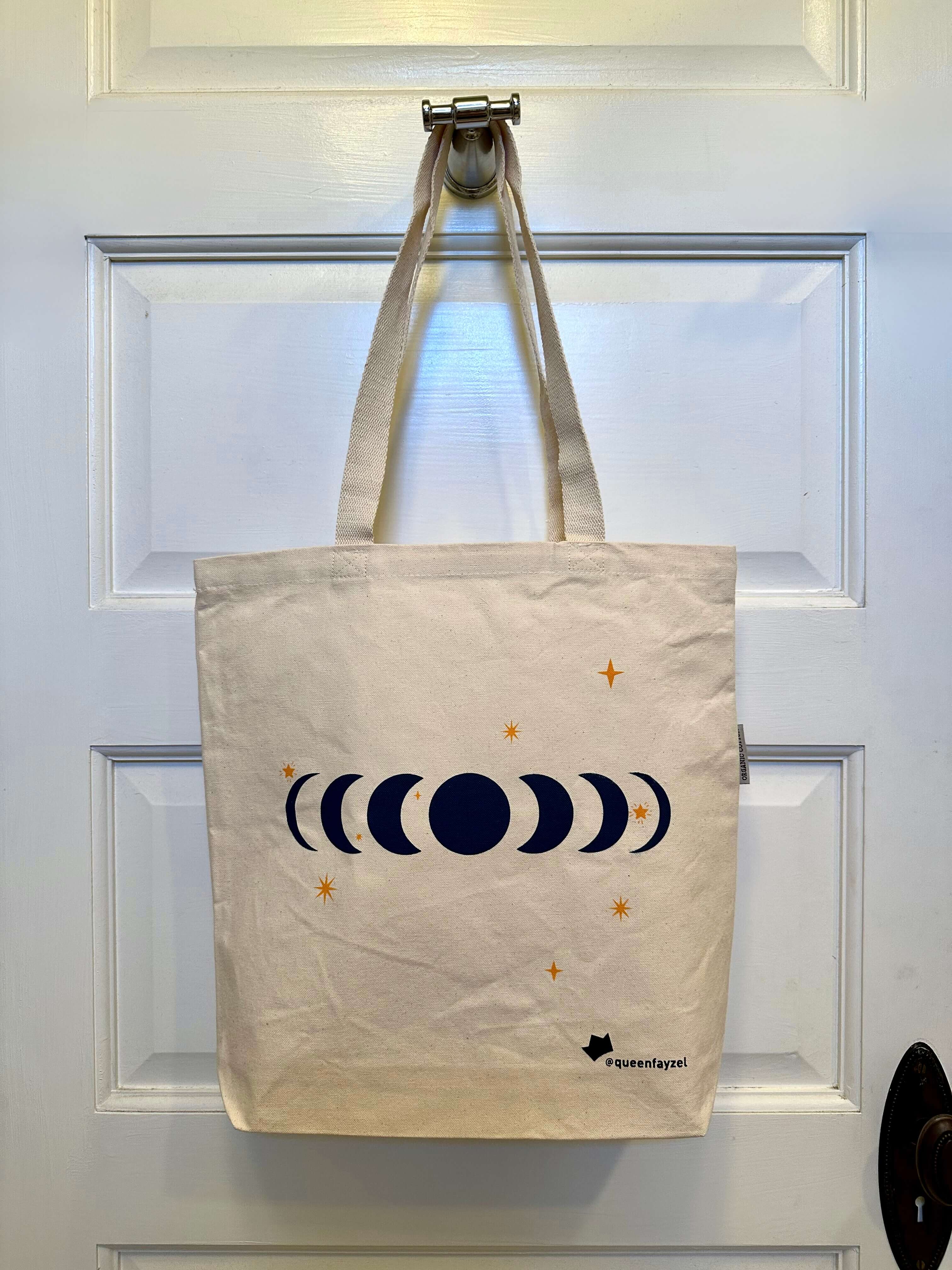 cotton tote bag with moon phases design hanging on a hook on the back of a white door.