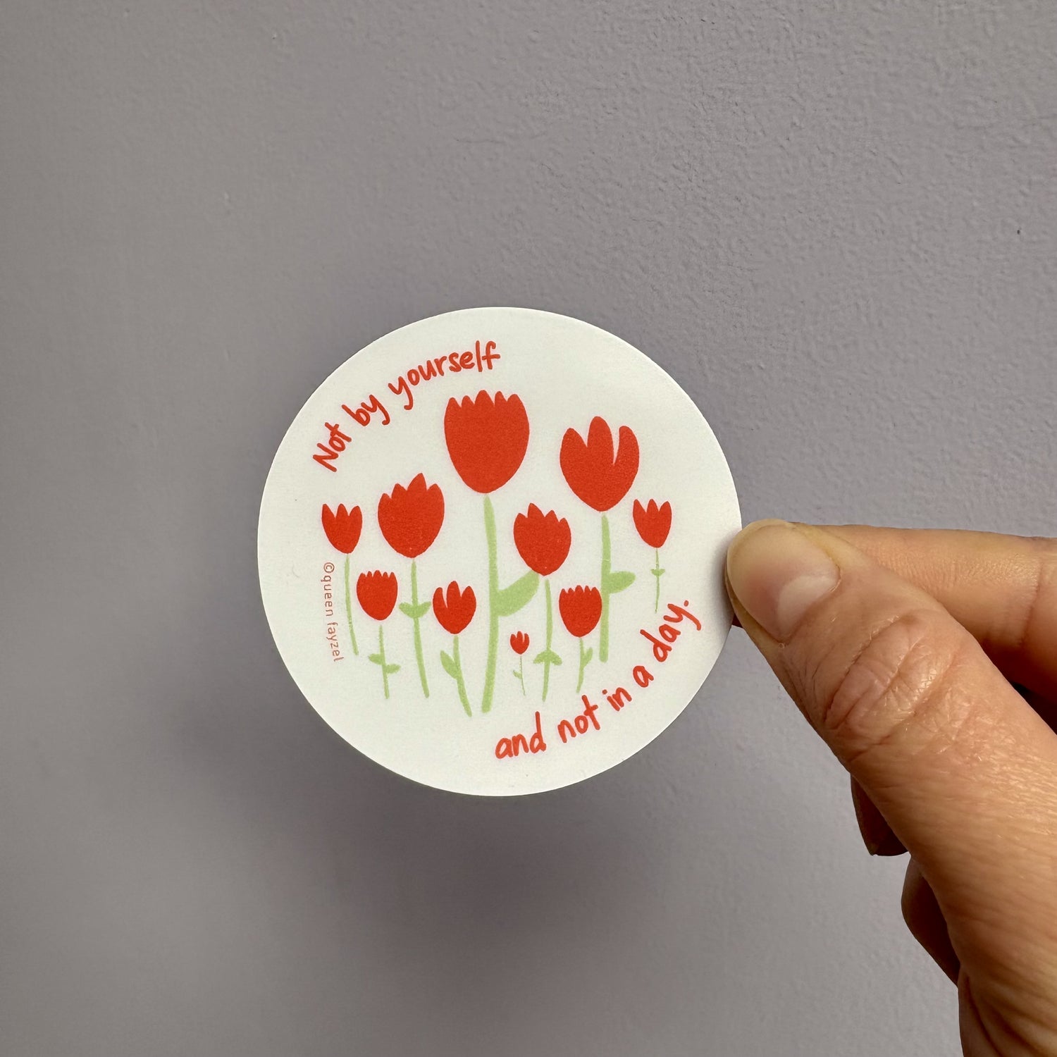 Round sticker with red tulips and text "not by yourself and not in a day" held by a hand against a gray background