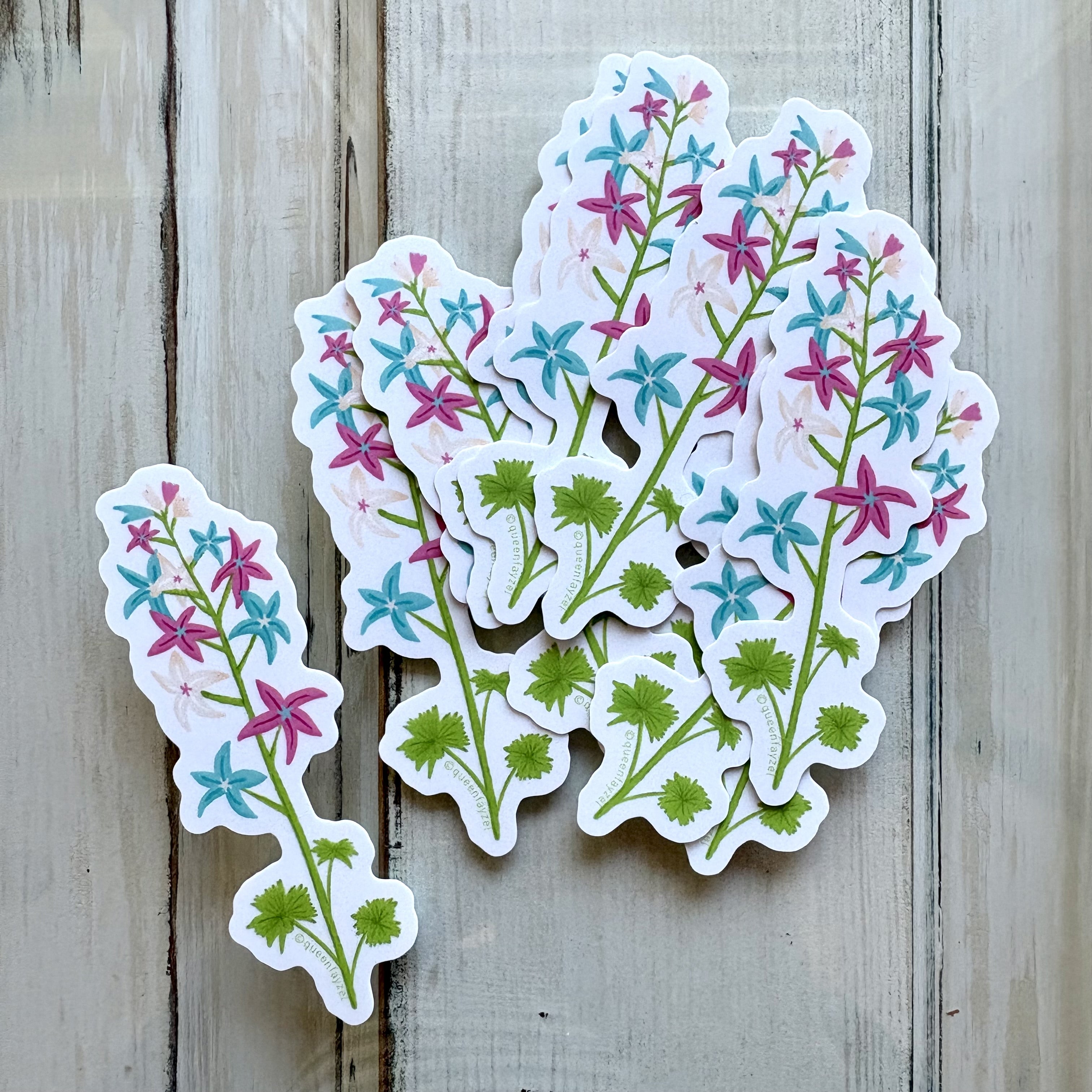 Artistic flower stalk stickers featuring the trans pride color scheme displayed on a light wooden background.