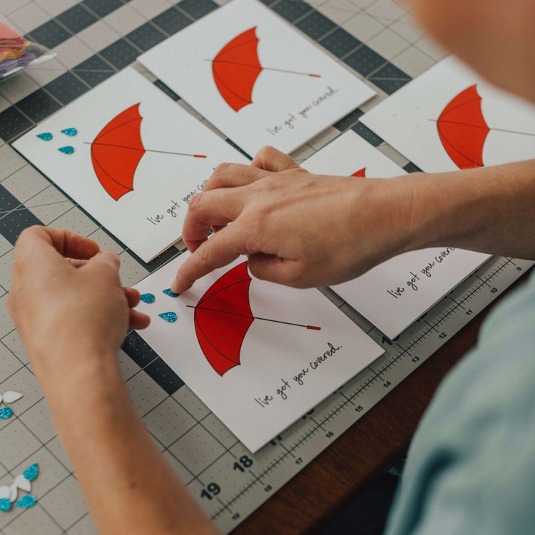 Photo showing a pair of hands putting small blue glitter raindrops on individual greeting cards