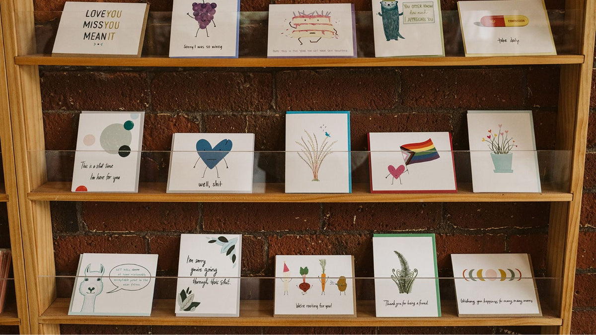 a selection of greeting cards with bright, colorful illustrations, sitting on wooden shelves against a brick background