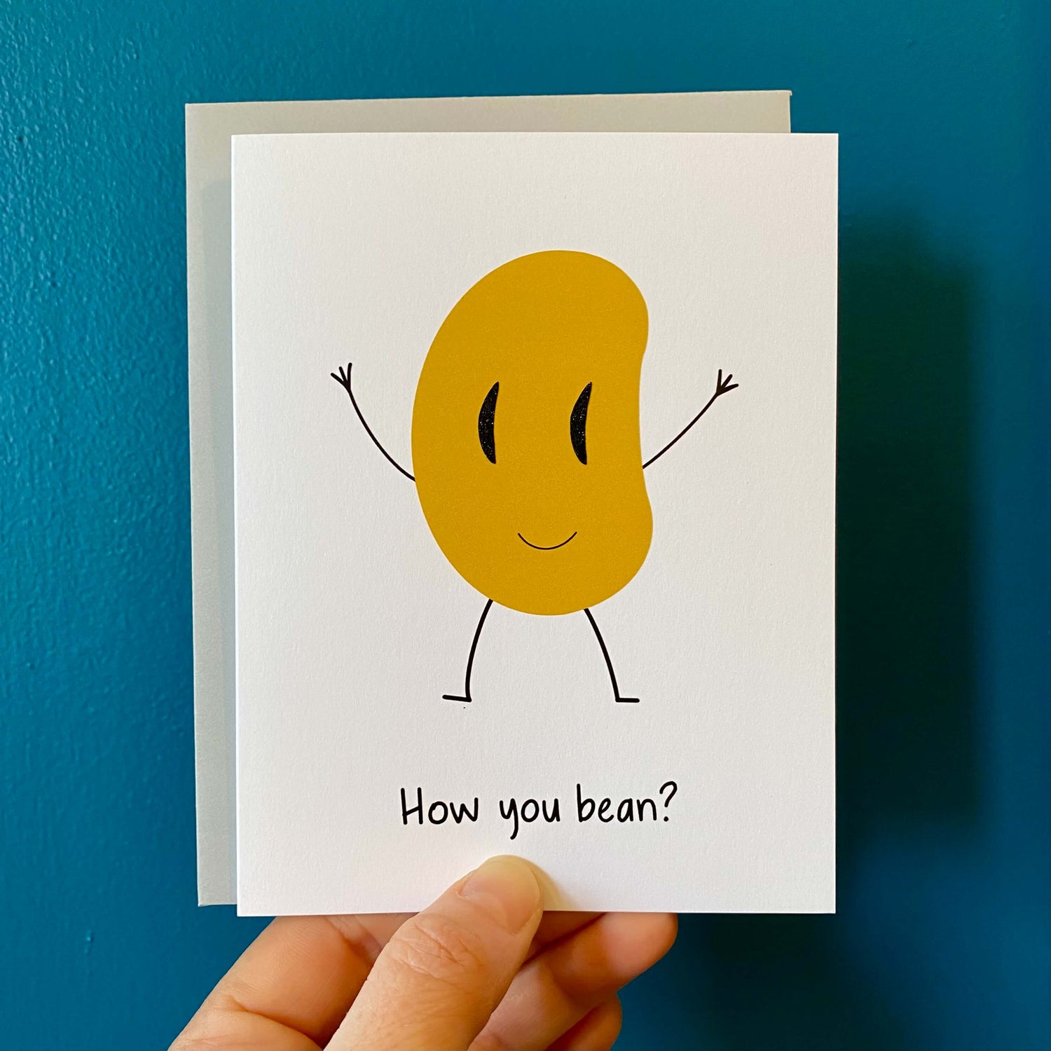 hand holding a funny greeting card with a waving bean and the text, &quot;How you bean?&quot;