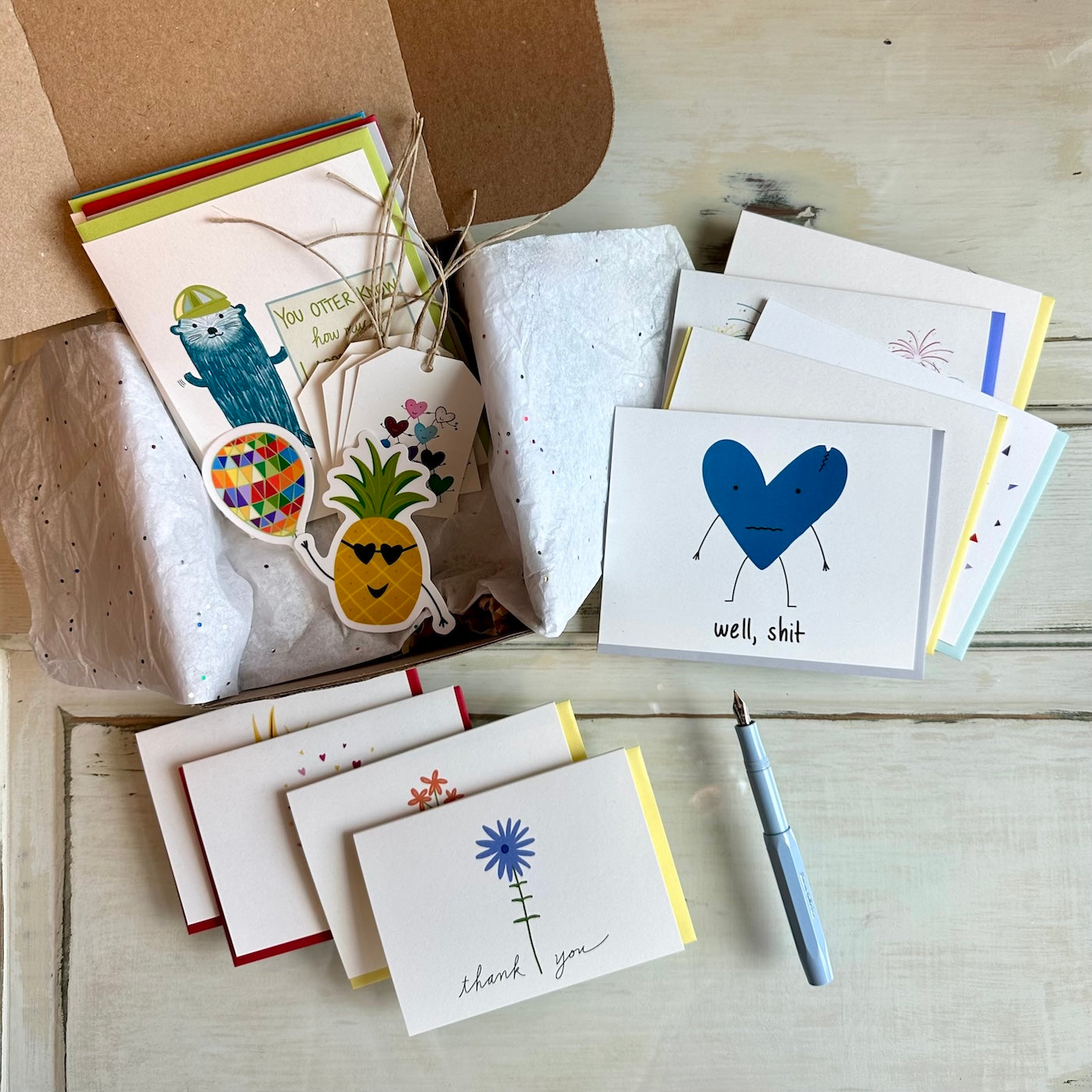 A gift box set of greeting cards, stickers, and gift tags against a white wood background. There is a fountain pen next to the cards.