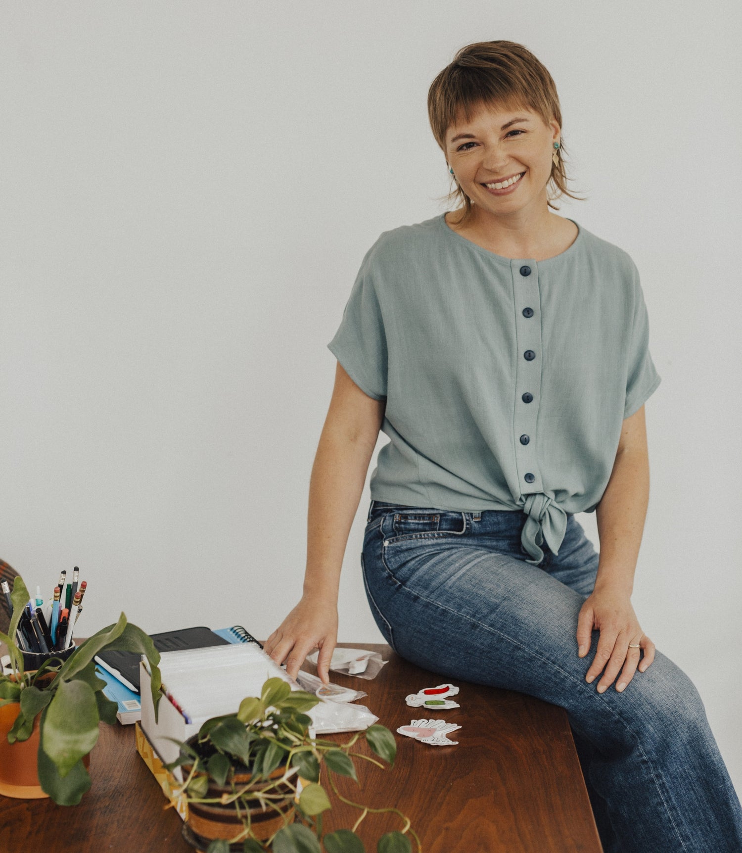 Faye sitting on a wooden desk. Beside her are some of her handmade greeting cards, a pen holder with pens, and leaves overflowing from a green plant