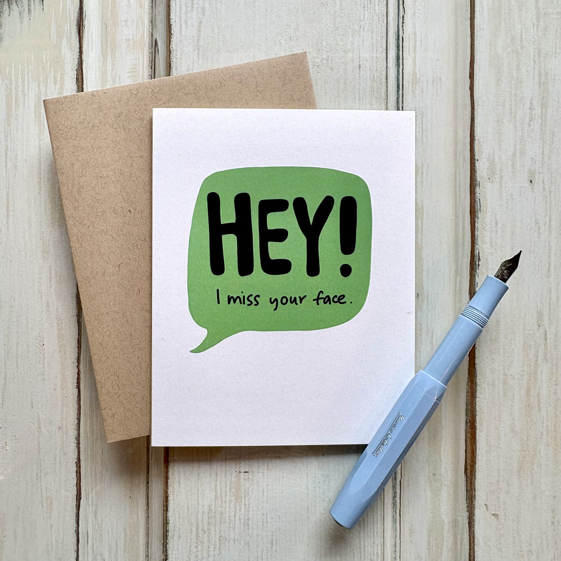 funny friendship greeting card with a green speech bubble and the text "HEY! I miss your face" paired with a kraft envelope and shown with a fountain pen on a wooden desk.