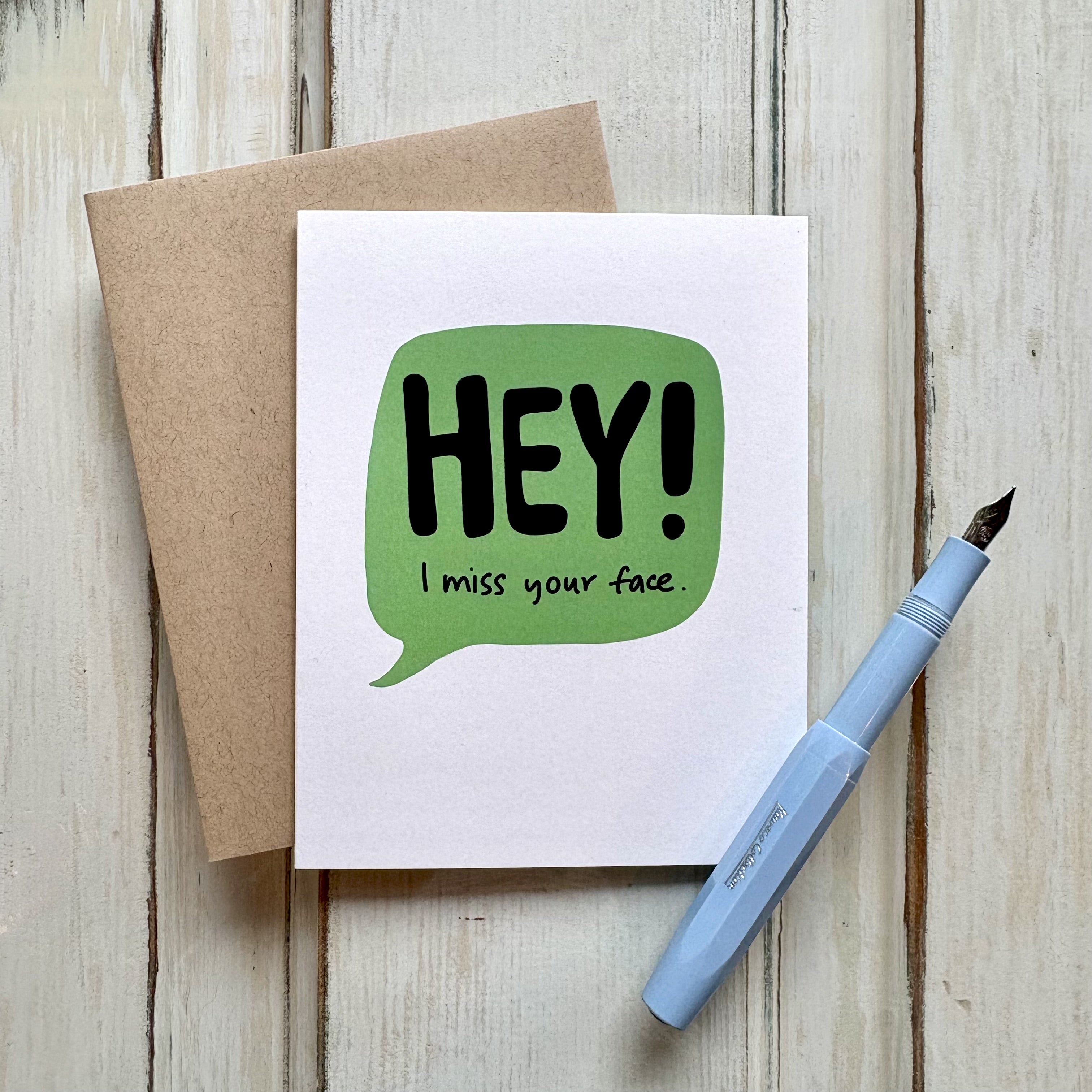 funny friendship greeting card with a green speech bubble and the text "HEY! I miss your face" paired with a kraft envelope and shown with a fountain pen on a wooden desk.