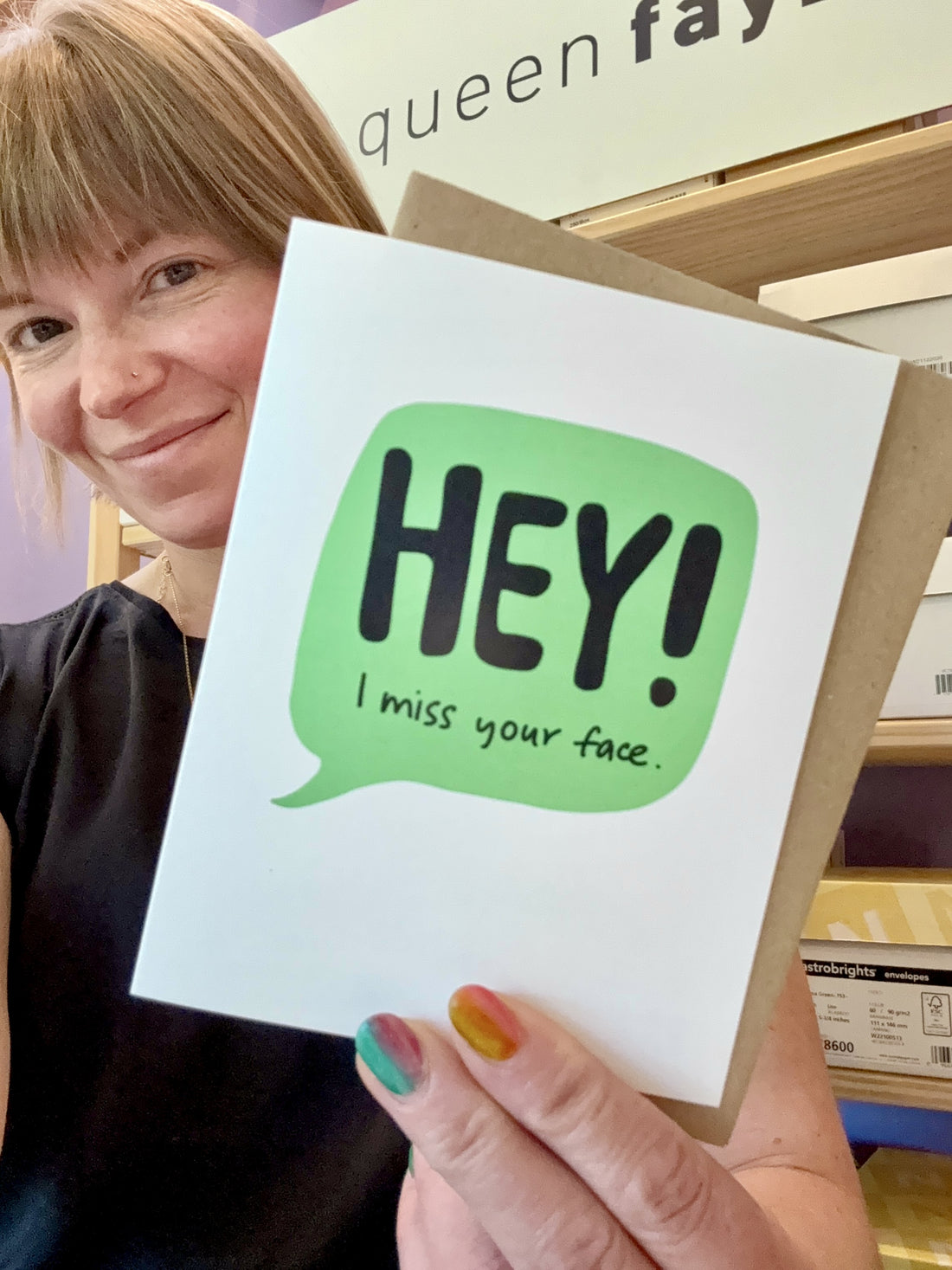 a white woman holding a friendship greeting card up to the camera. the card is white with a green speech bubble and the words, "HEY! I miss your face."