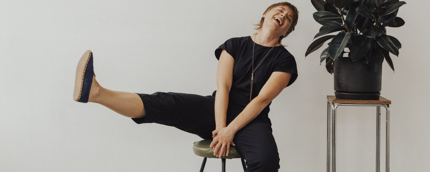 Faye, a greeting card designer and maker, sitting on a stool kicking one leg back and laughing.