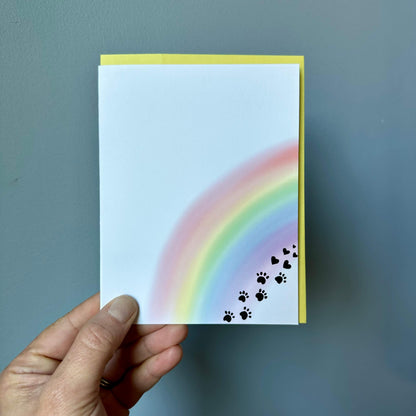 hand holding a pet loss sympathy card against a gray background. the card has a watercolor rainbow with black paw prints that transition to hearts.