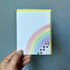 hand holding a pet loss sympathy card against a gray background. the card has a watercolor rainbow with black paw prints that transition to hearts.