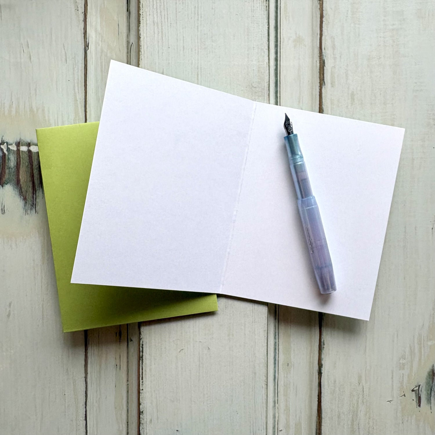 Open blank greeting card with a pen and bright green envelope on a wooden surface.