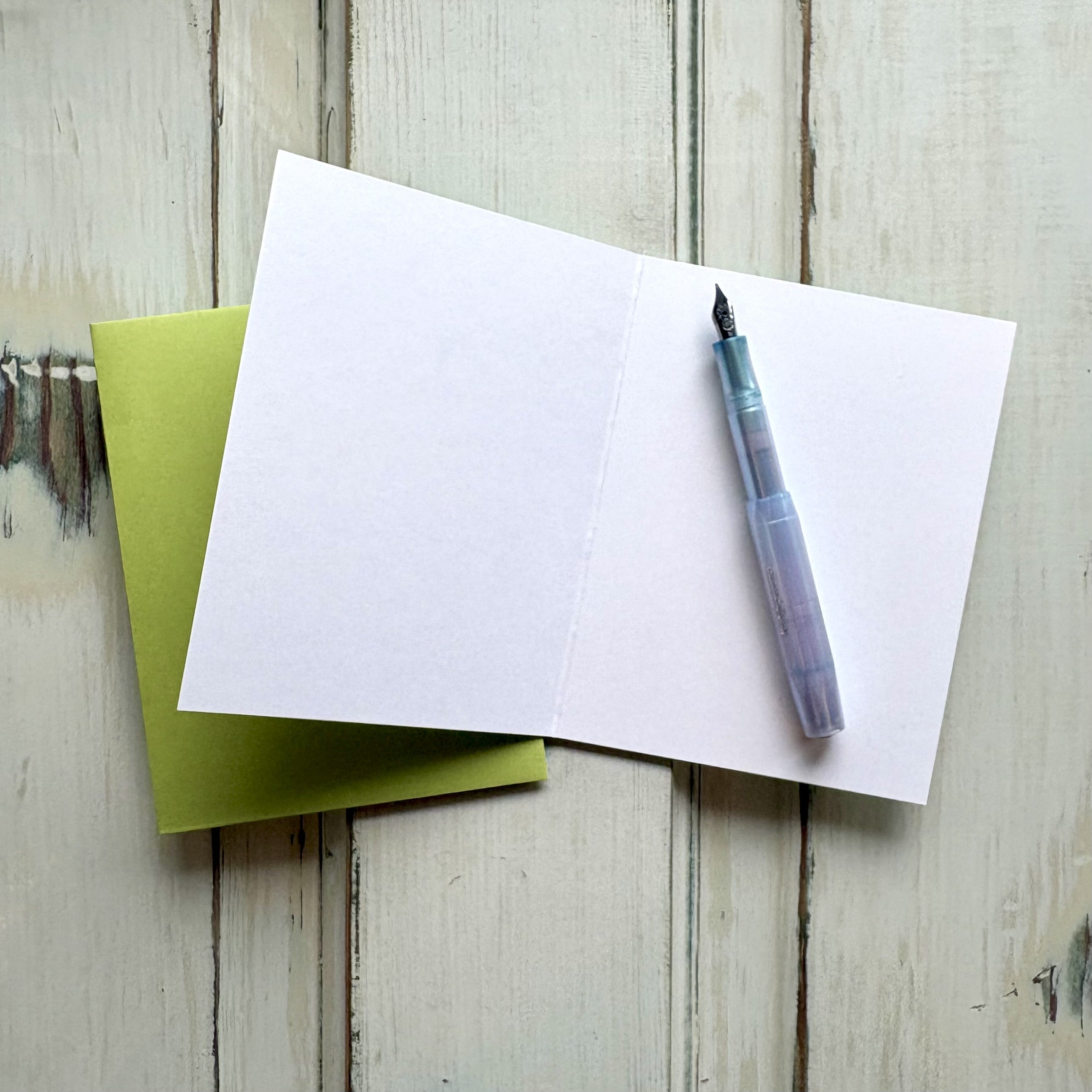 Open blank greeting card with a pen and bright green envelope on a wooden surface.