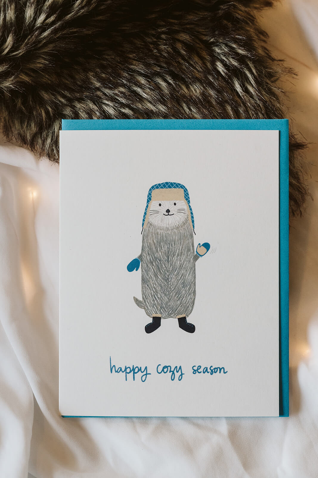 seasonal greeting card with a cozy otter in a flap hat and mittens