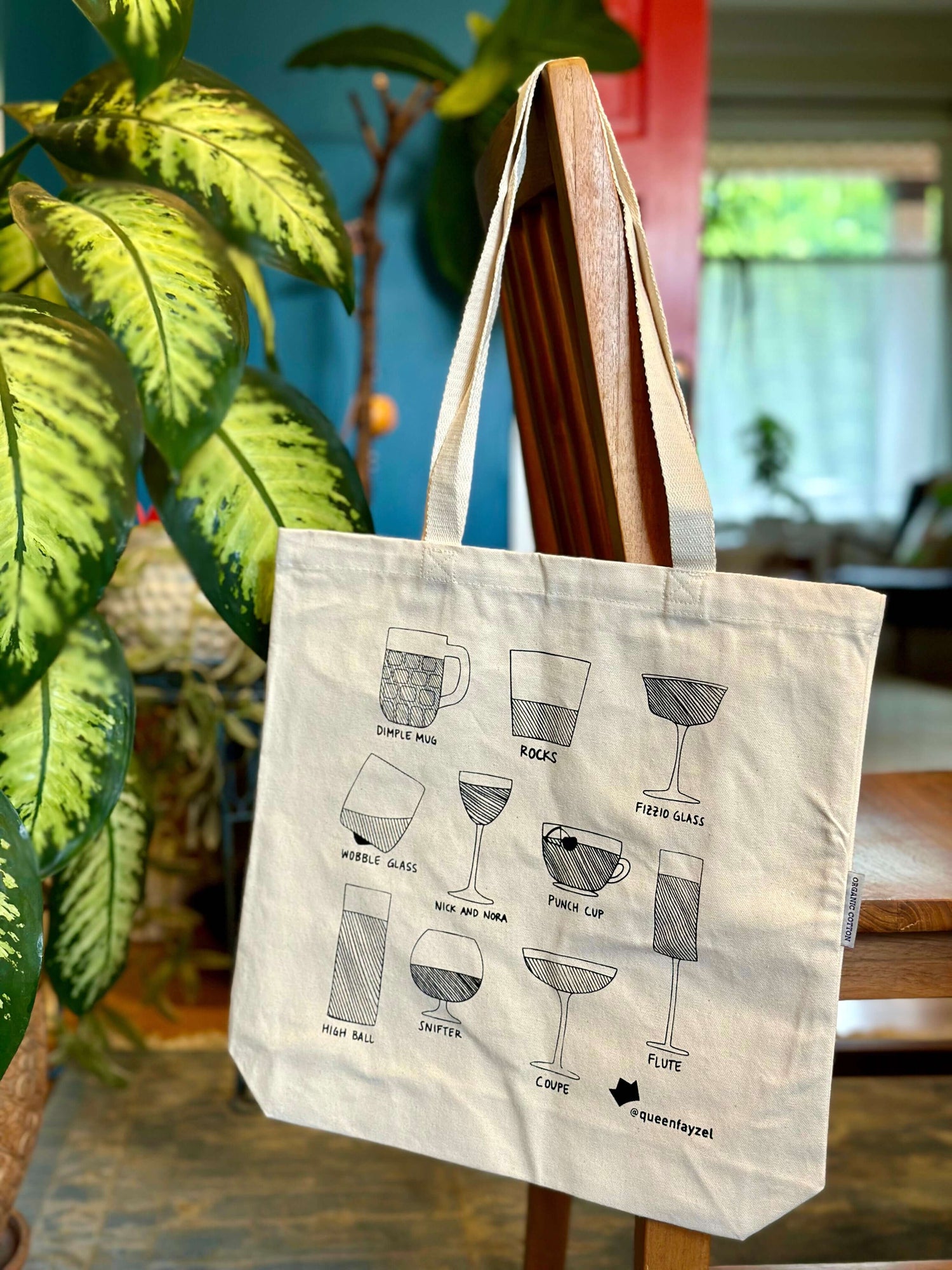 A white tote bag with a printed drink glass design, hanging on a wooden chair with a blue wall and plants in the background.