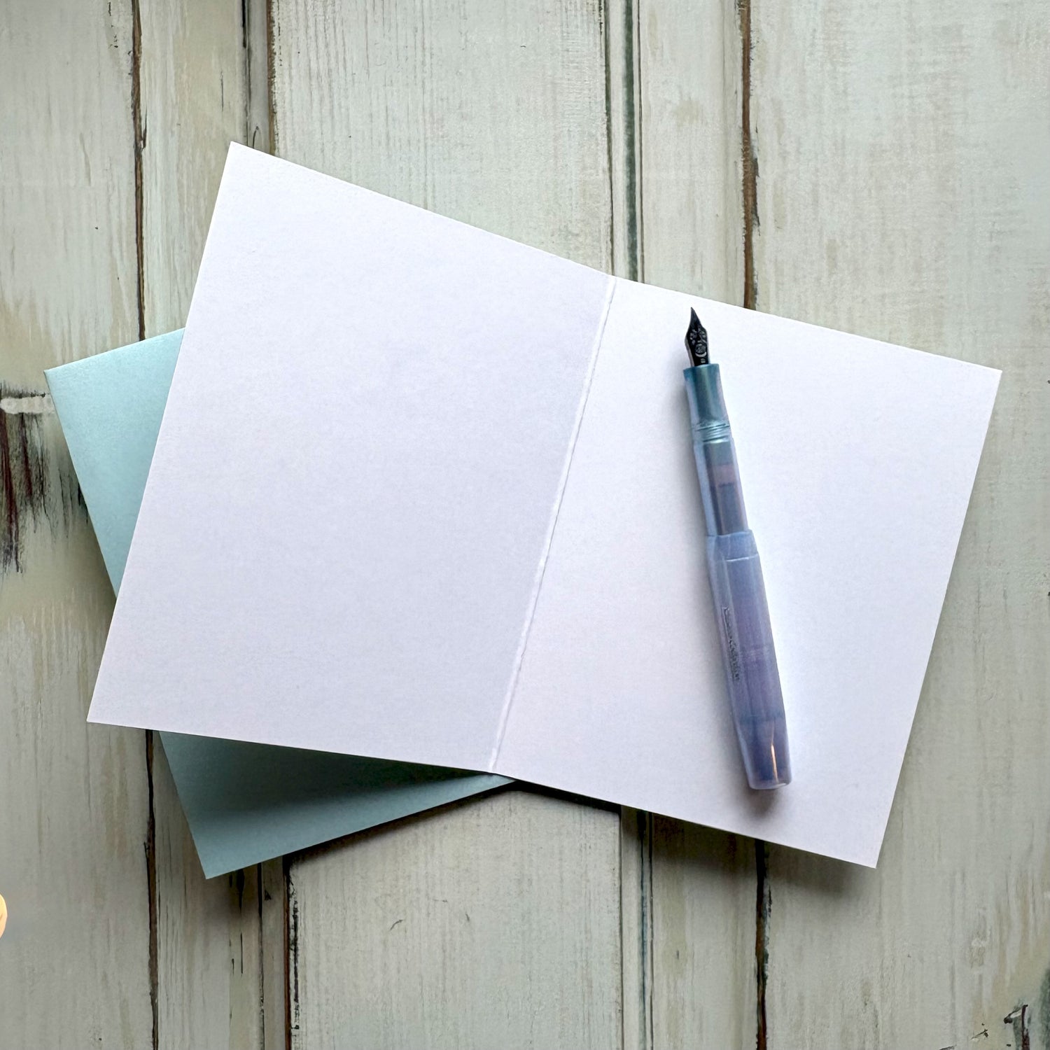 Open blank greeting card with a fountain pen and light blue envelope on a wooden surface.