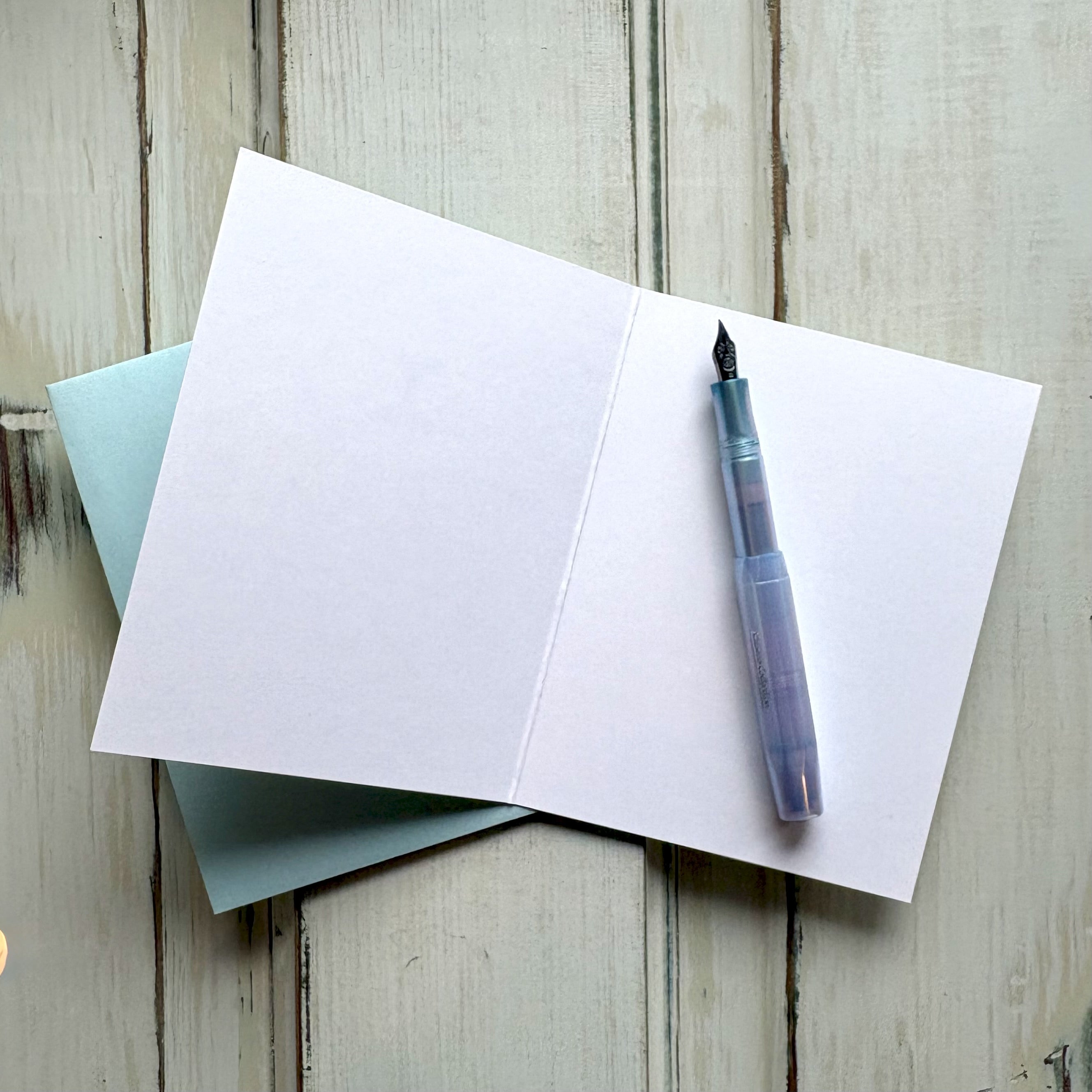 Open blank greeting card with a fountain pen and light blue envelope on a wooden surface.