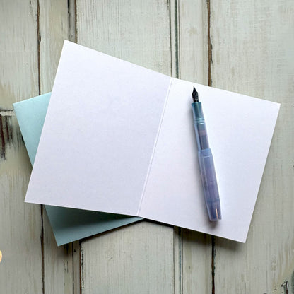 Open blank greeting card with a fountain pen and light blue envelope on a wooden surface.