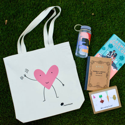 cute tote bag with waving heart design laid out on grass with a water bottle, journal, book, and set of greeting cards