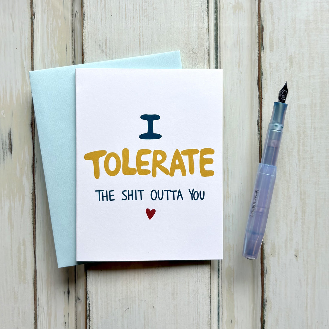 A funny anniversary card in a light blue envelope featuring "I TOLERATE THE SHIT OUTTA YOU" in playful blue and yellow fonts with a red heart.