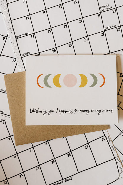 congratulations card with moon phase design and the words, "wishing you happiness for many, many moons" against a background of calendar pages.