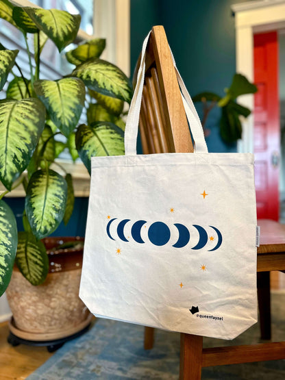 cotton tote bag with moon phase design hanging on the back of a wooden chair with a plant and blue wall in the background