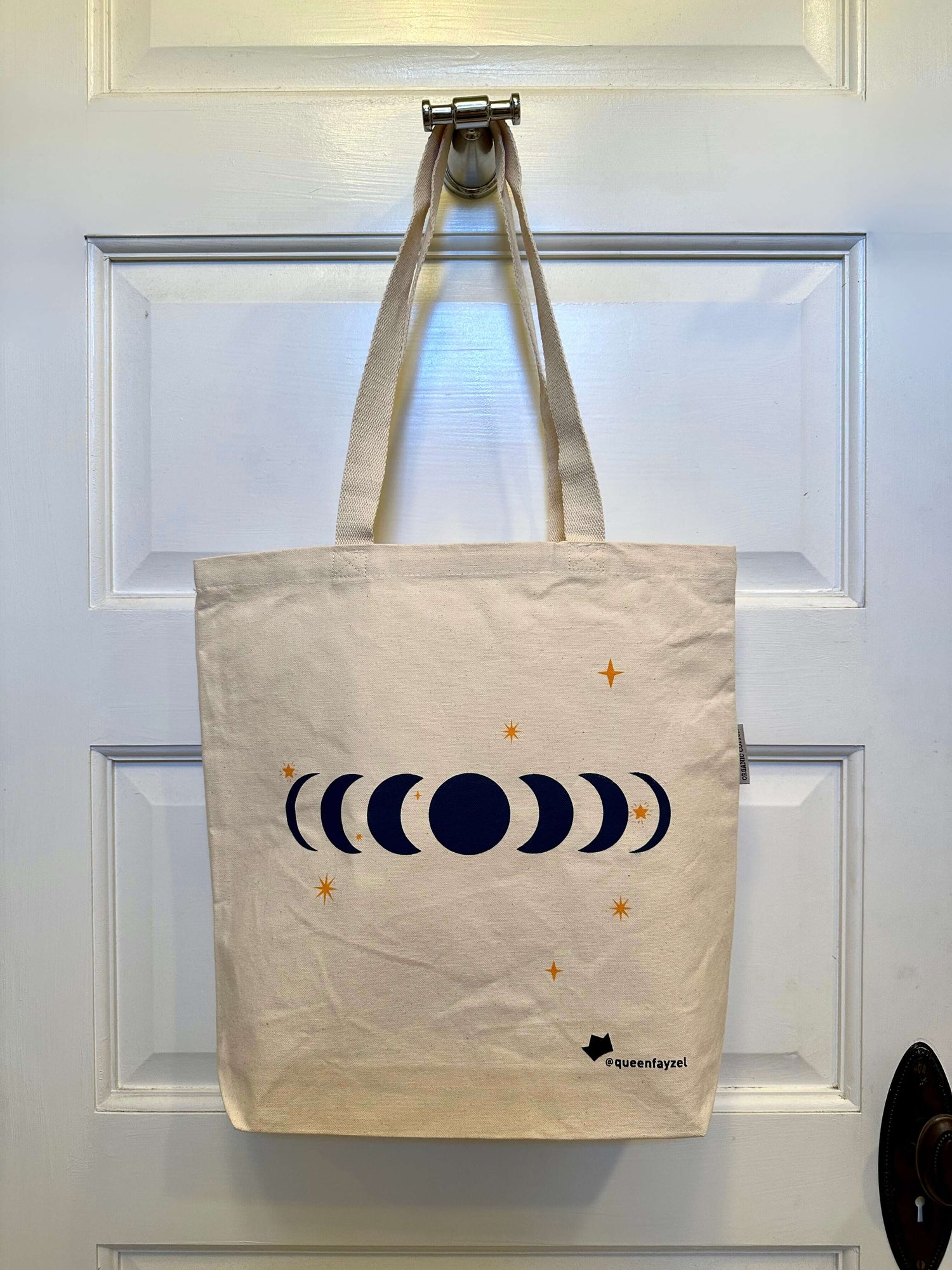 cotton tote bag with moon phases design hanging on a hook on the back of a white door.