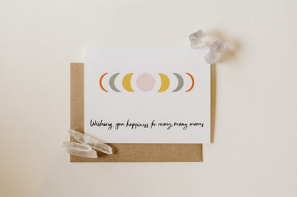 a wedding congratulations card with moon phases illustration and text reading "wishing you happiness for many, many moons" the card is against a cream background, with crystals holding down two corners of the card, and the kraft envelope.
