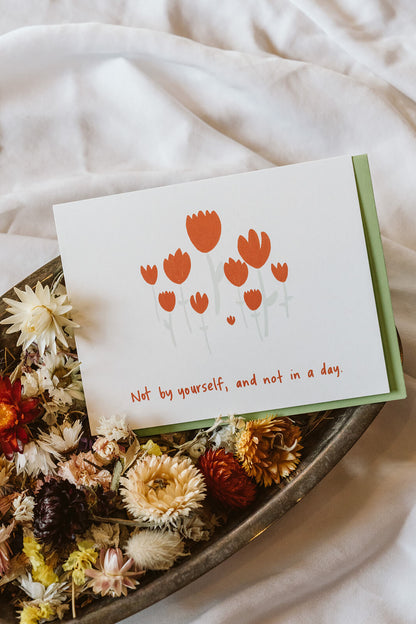 An encouragement affirmation card reading"Not by yourself, and not in a day," sits nestled in a bowl of dried flowers.