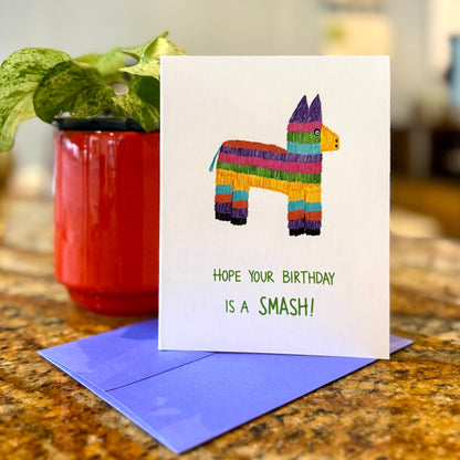 Birthday card with colorful llama pinata design and purple envelope on a table.