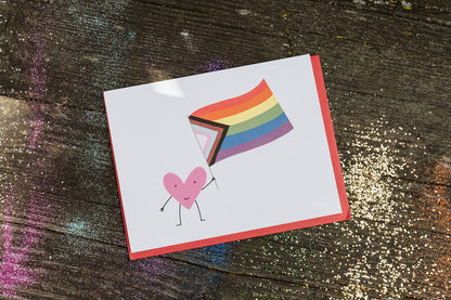 greeting card celebrating the queer community with a smiling heart waving a progress pride flag