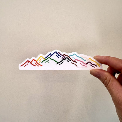 A hand holds a colorful rainbow sticker featuring a mountain range designed in the colors of the intersex progress flag.