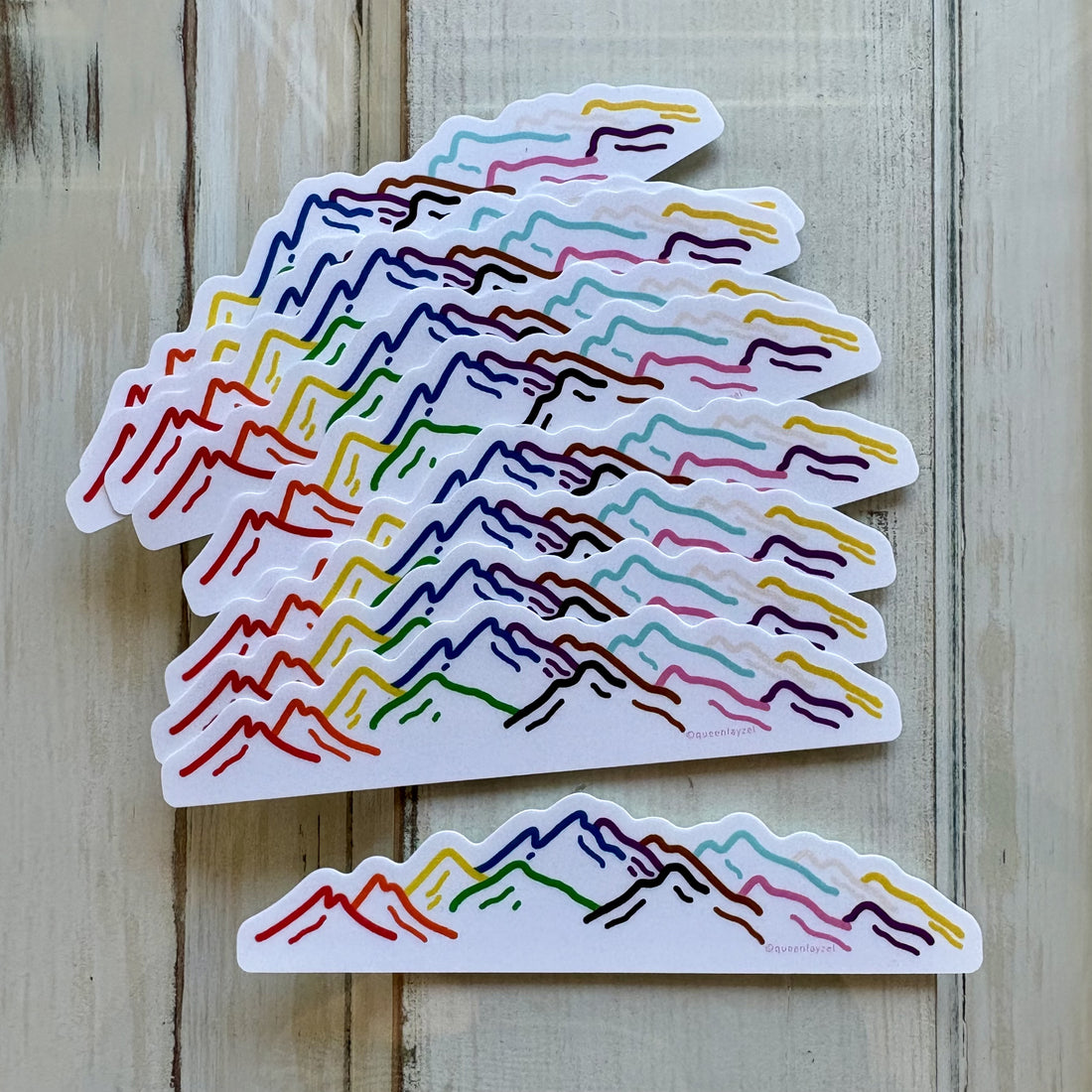 Scattered pride stickers showing mountain silhouettes in intersex progress pride colors on a rustic wood surface.