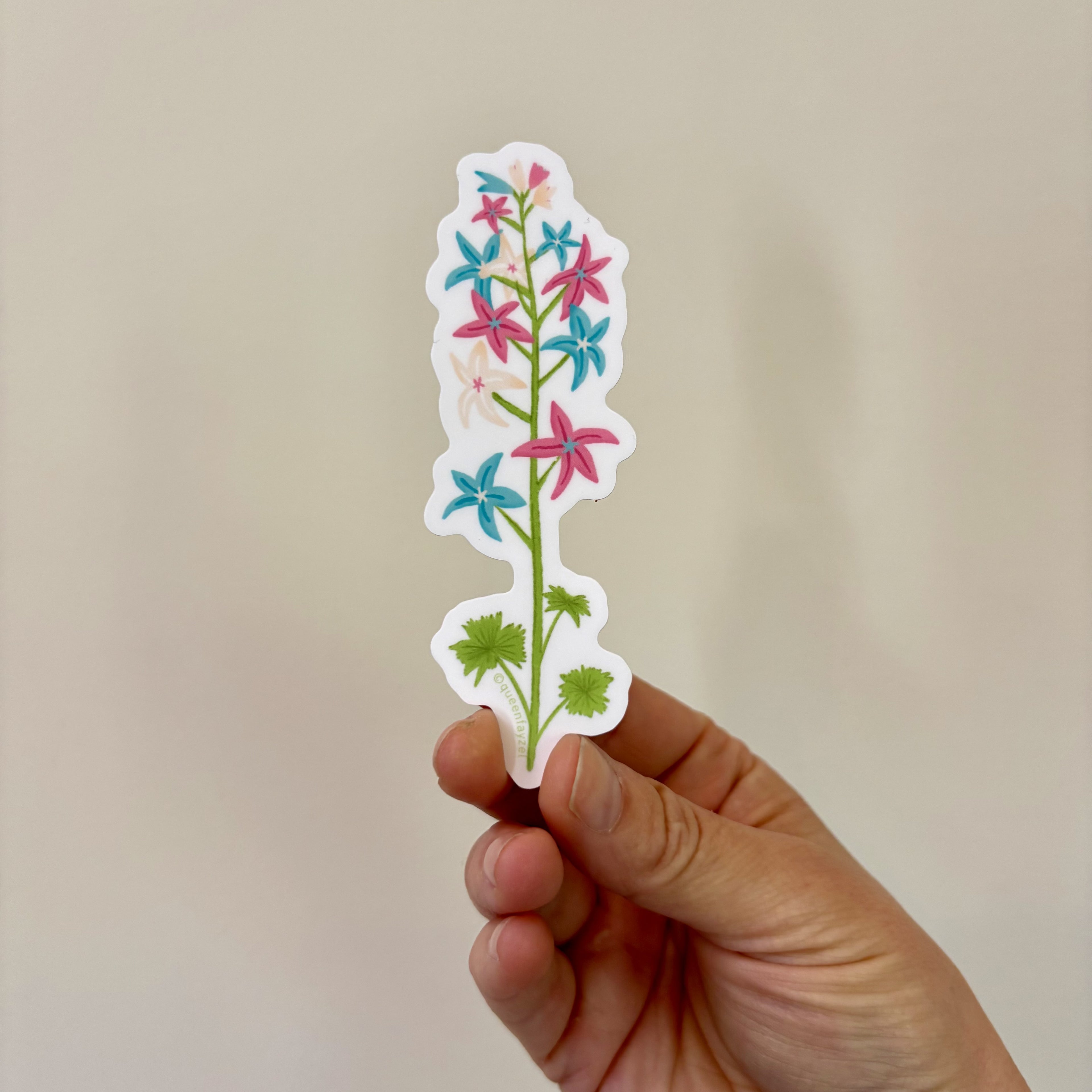 A sticker of a flower stalk with pink, blue, and white colors inspired by the trans pride flag, held in a hand.