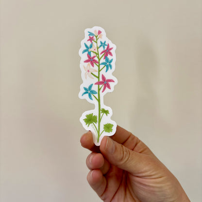 A sticker of a flower stalk with pink, blue, and white colors inspired by the trans pride flag, held in a hand.