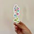 A sticker of a flower stalk with pink, blue, and white colors inspired by the trans pride flag, held in a hand.