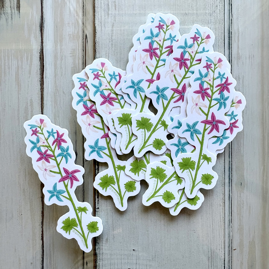 Artistic flower stalk stickers featuring the trans pride color scheme displayed on a light wooden background.