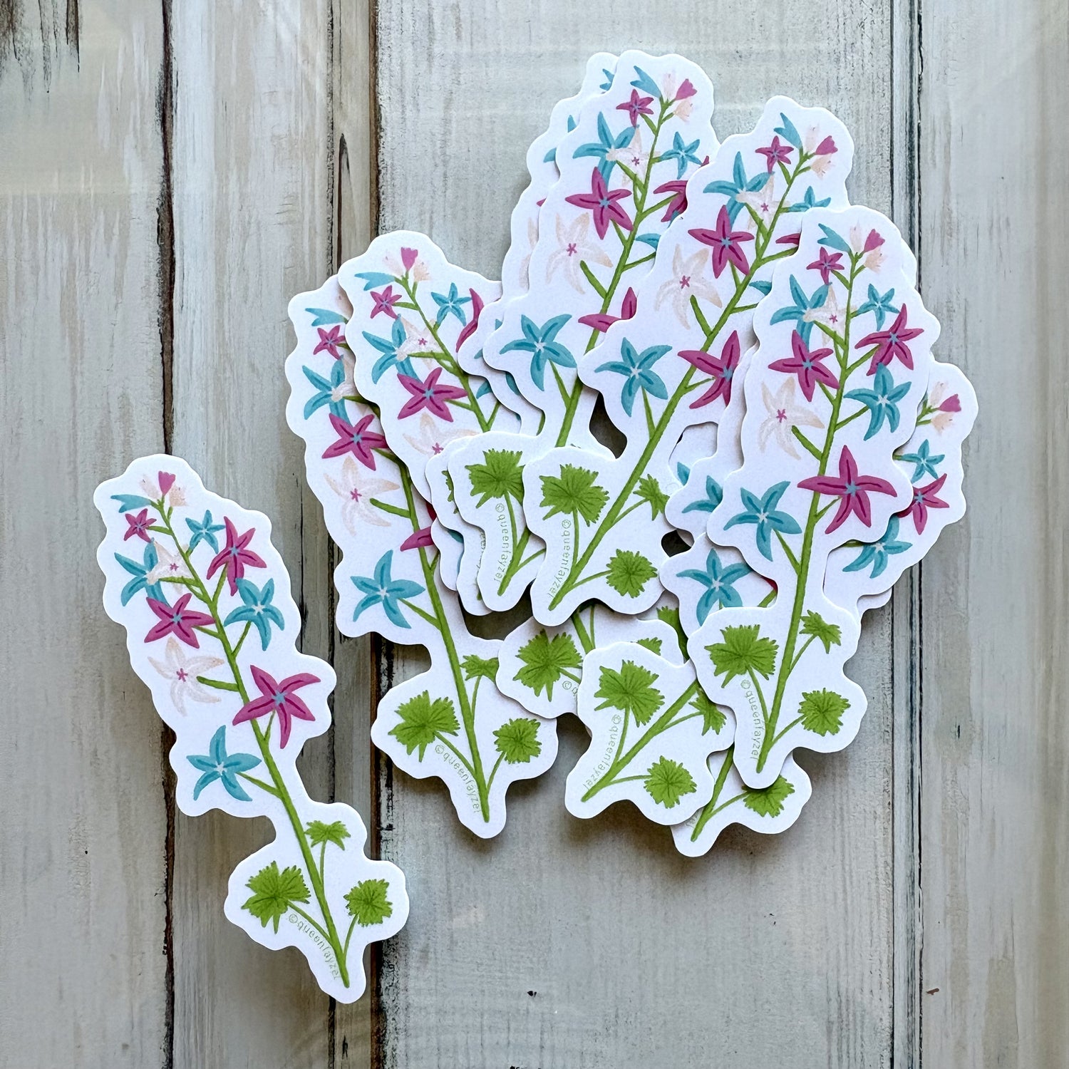Artistic flower stalk stickers featuring the trans pride color scheme displayed on a light wooden background.