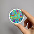 Round sticker with brightly colored ink-blob designs and yellow text reading "Trust yourself" held by a hand against a gray background.