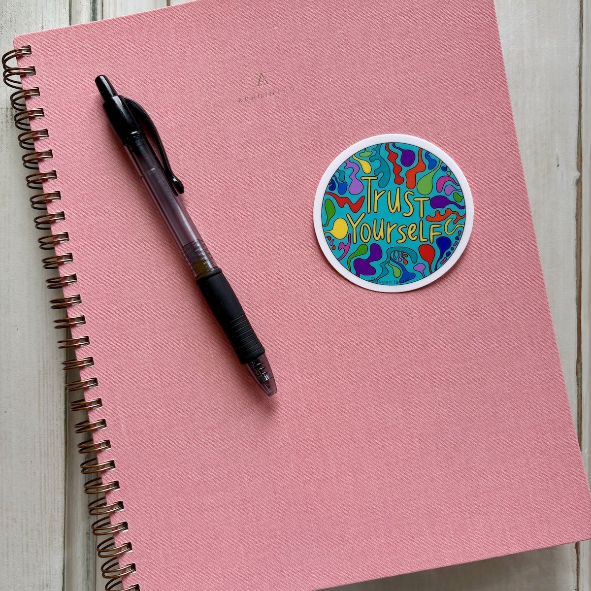 Pink notebook with a pen and a colorful &quot;Trust yourself&quot; sticker on a light wooden surface