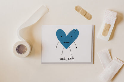 a funny sympathy card with a cracked blue heart and the words "well, shit." the card is agains a white background with bandages arranged around it.