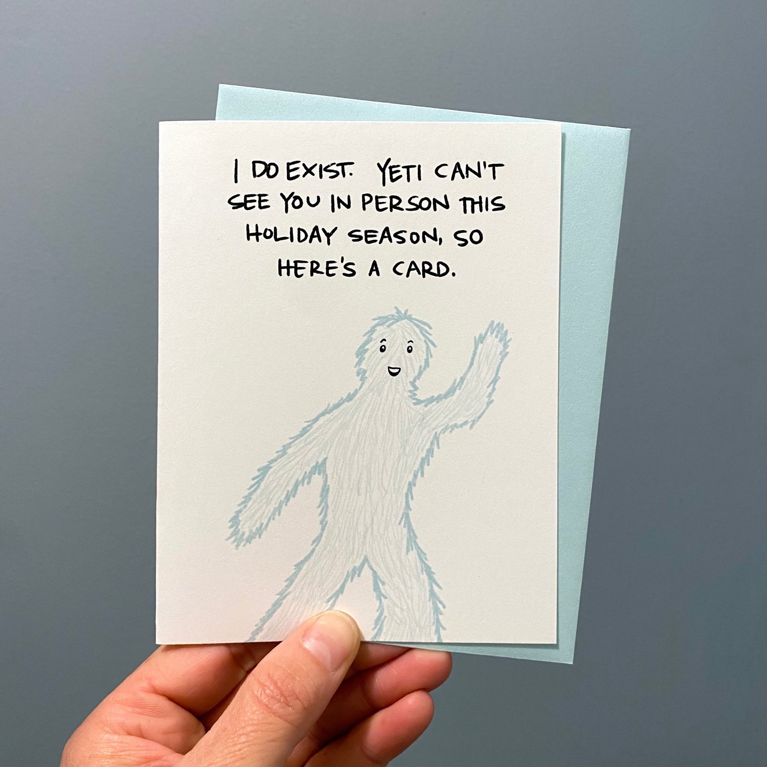 Image of a hand holding a funny holiday card with a waving yeti.
