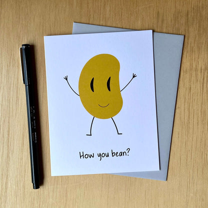 Punny, funny greeting card with a smiling bean and the words, &quot;How you bean?&quot; The card is next to a gray envelope and black pen.